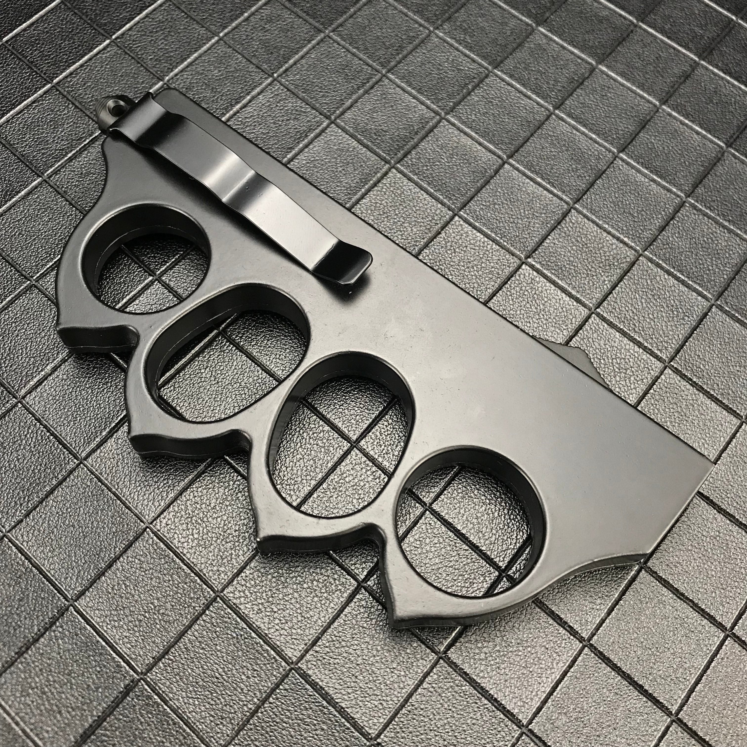 The back side of a closed black 1918 U.S. knuckle duster OTF knife, showing the silver pocket clip and four finger holes on a dark grid background.