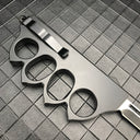 The back side of a closed black 1918 U.S. knuckle duster OTF knife, showing the silver pocket clip and glass breaker on a grid background.