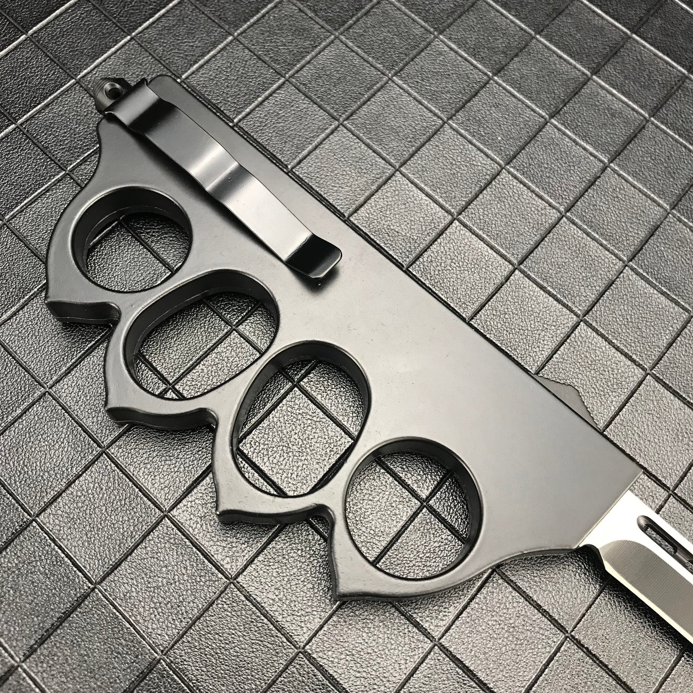 The back side of a closed black 1918 U.S. knuckle duster OTF knife, showing the silver pocket clip and glass breaker on a grid background.