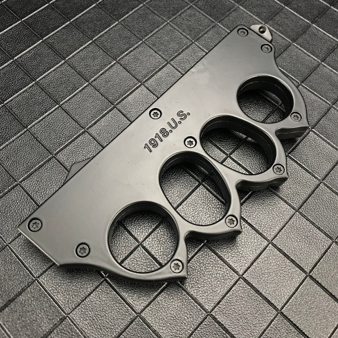 A close-up of a closed black knuckle duster OTF knife, showing the engraved "1918 U.S." text and screw details on a dark grid background.
