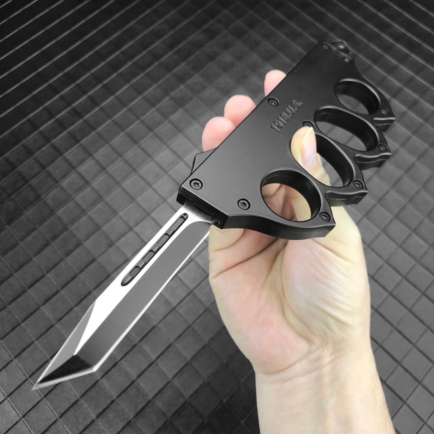 A hand holding an open 1918 U.S. style black knuckle duster OTF knife with a silver tanto blade, displayed on a dark grid background.