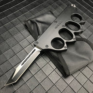 An open black 1918 U.S. knuckle duster OTF knife resting on its black nylon sheath, displayed on a dark grid background.
