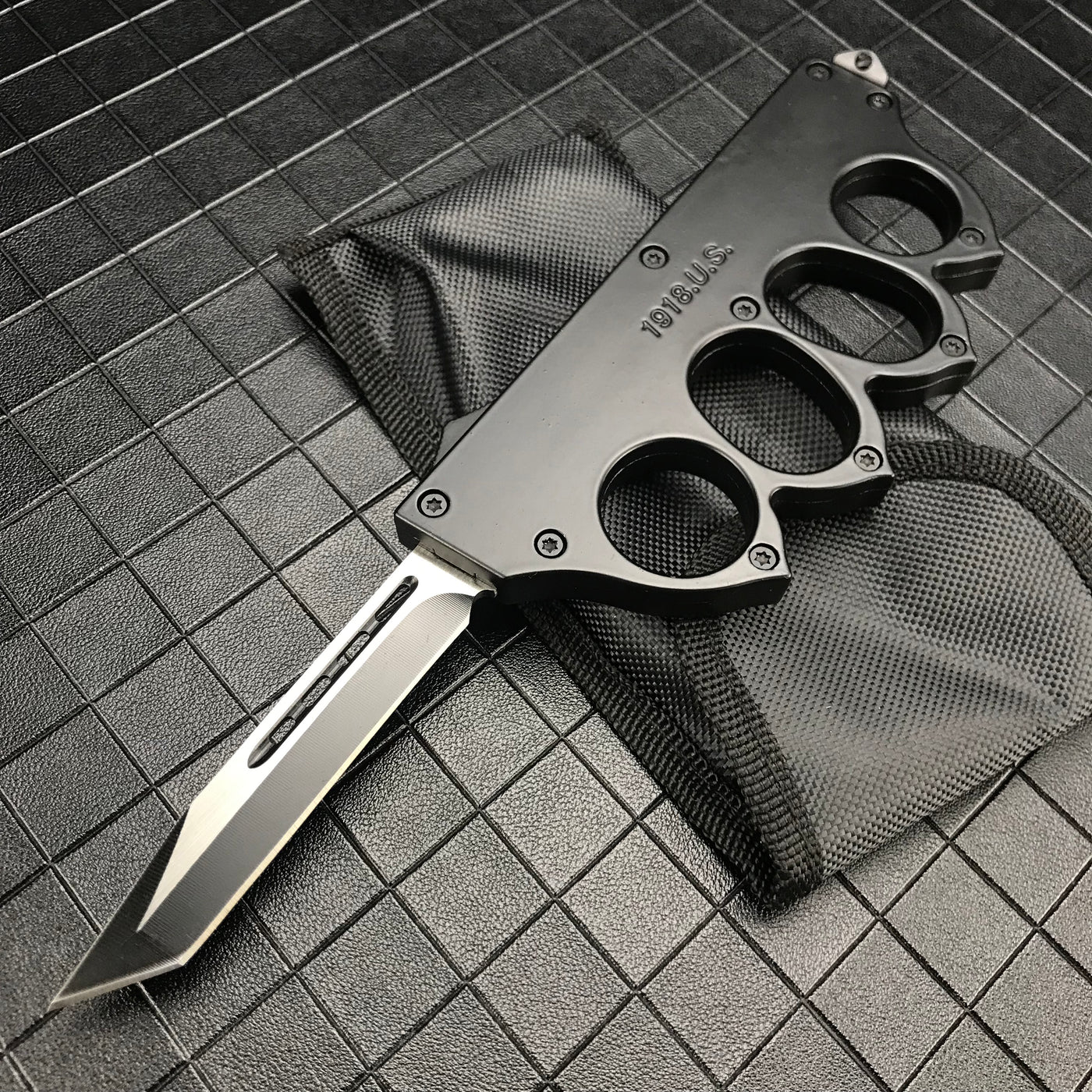An open black 1918 U.S. knuckle duster OTF knife resting on its black nylon sheath, displayed on a dark grid background.