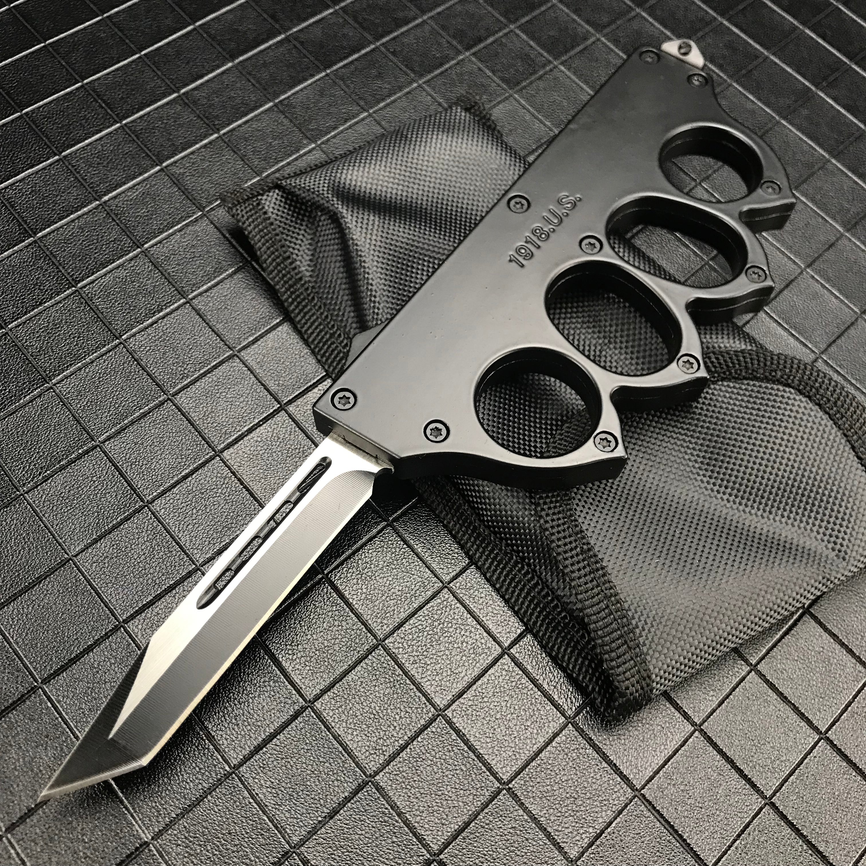 An open black 1918 U.S. knuckle duster OTF knife resting on its black nylon sheath, displayed on a dark grid background.