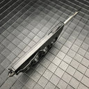 A side view of a closed black OTF knife with a knuckle duster handle, highlighting the side-mounted firing button and glass breaker tip on a grid background.