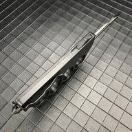 A side view of a closed black OTF knife with a knuckle duster handle, highlighting the side-mounted firing button and glass breaker tip on a grid background.