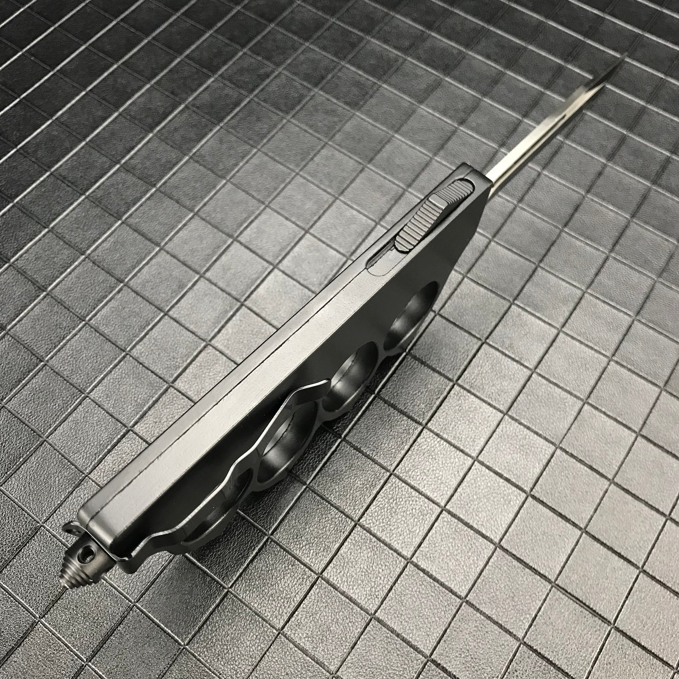 A side view of a closed black OTF knife with a knuckle duster handle, highlighting the side-mounted firing button and glass breaker tip on a grid background.
