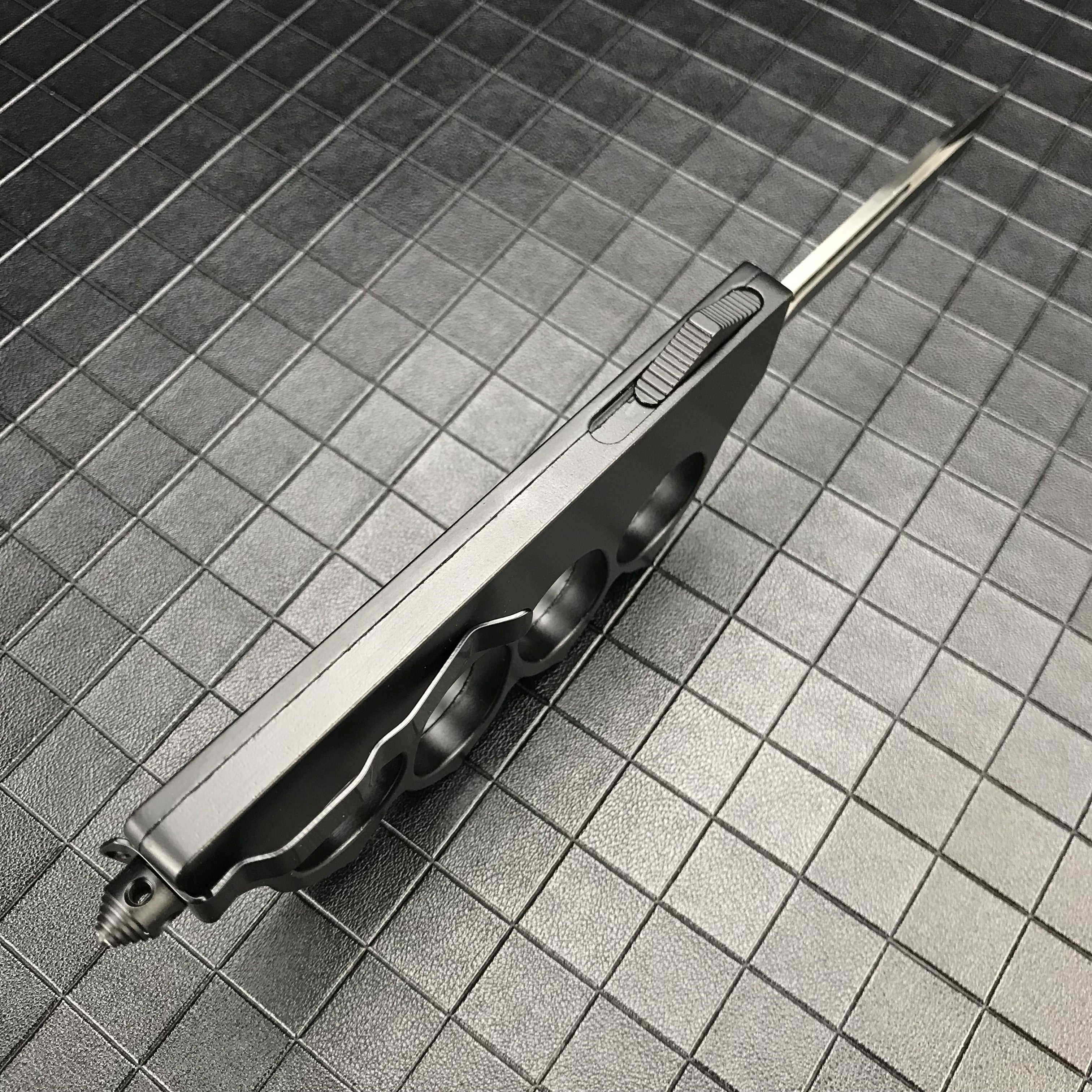A side view of a closed black OTF knife with a knuckle duster handle, highlighting the side-mounted firing button and glass breaker tip on a grid background.