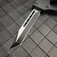 A close-up of the silver tanto blade of a 1918 U.S. OTF knife, showing the central groove and sharp tip on a dark grid background.