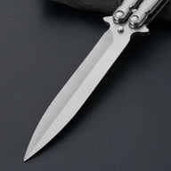 An extreme close-up of a balisong butterfly knife blade, showing the brushed stainless steel finish and sharp tip on a dark background.