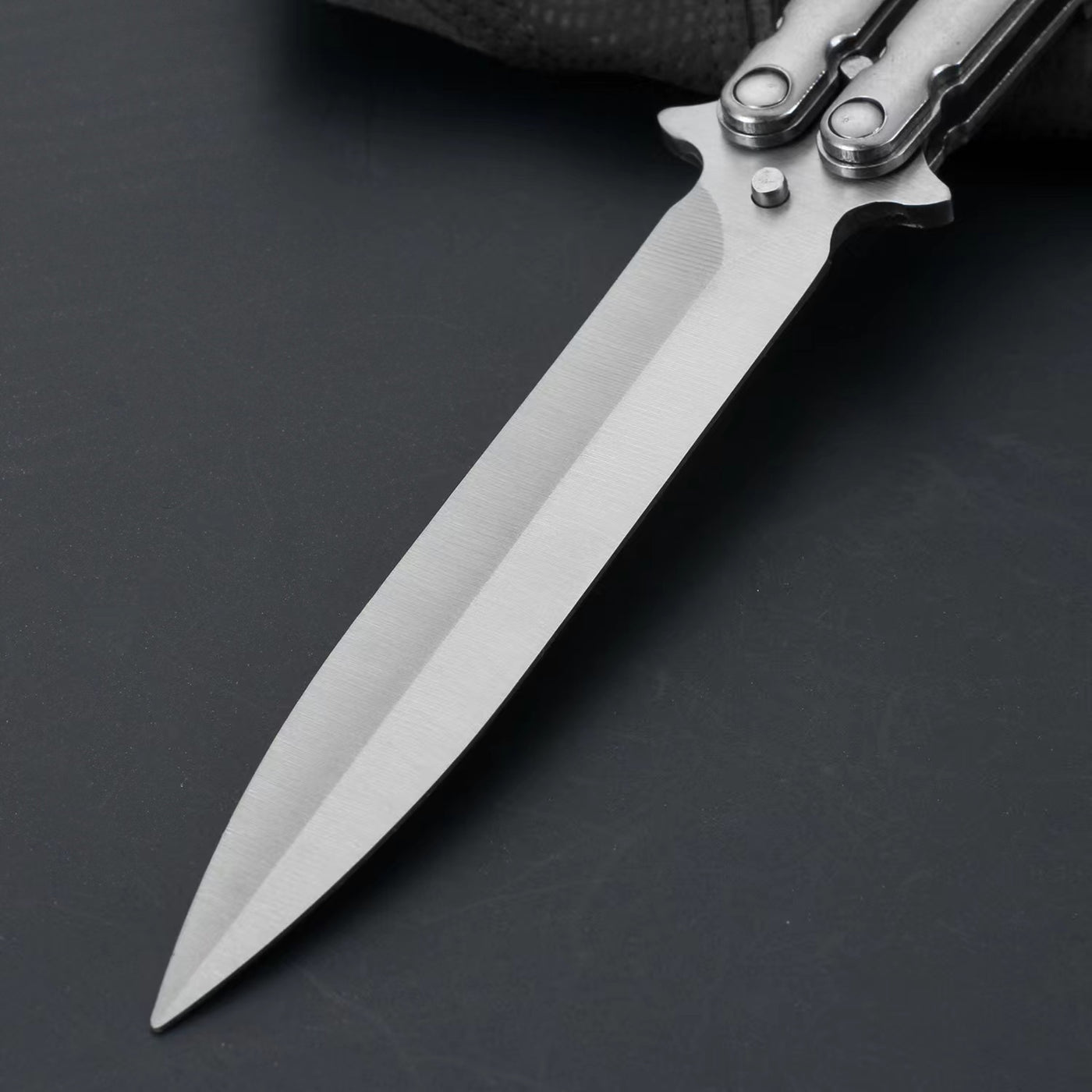 An extreme close-up of a balisong butterfly knife blade, showing the brushed stainless steel finish and sharp tip on a dark background.