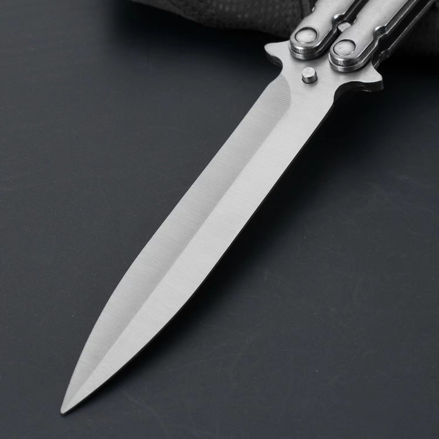 An extreme close-up of a balisong butterfly knife blade, showing the brushed stainless steel finish and sharp tip on a dark background.