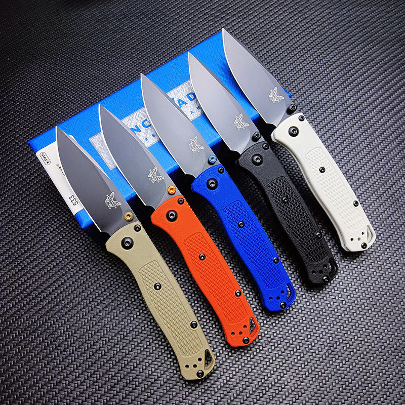 Five open Benchmade folding knives with tan, orange, blue, black, and white handles, resting on a blue Benchmade box on a dark textured surface.