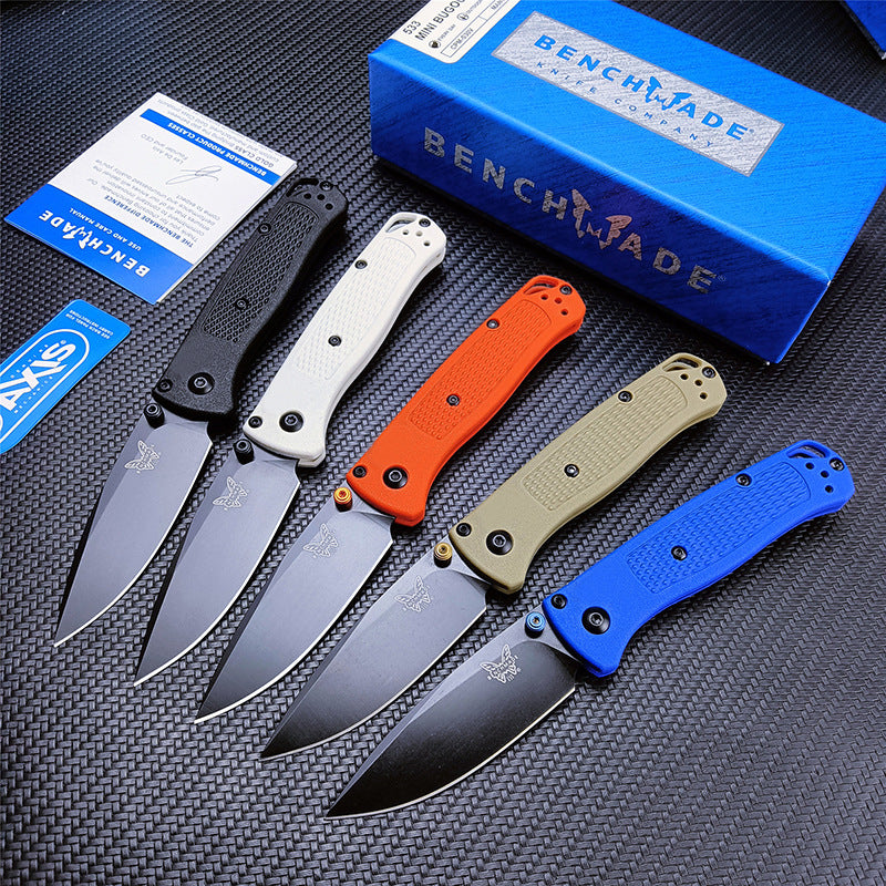 Five Benchmade folding knives in black, white, blue, orange, and tan, displayed with their blue box, warranty card, and sticker on a dark textured surface.
