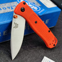 An open Benchmade folding knife with an orange handle, silver blade, and orange thumb stud, displayed with its blue box.