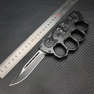 A black knuckle duster folding knife with a silver blade, featuring an embossed skull and motorcycle design with the text "BIKER USA", displayed with a metal ruler on a dark textured background for size reference.