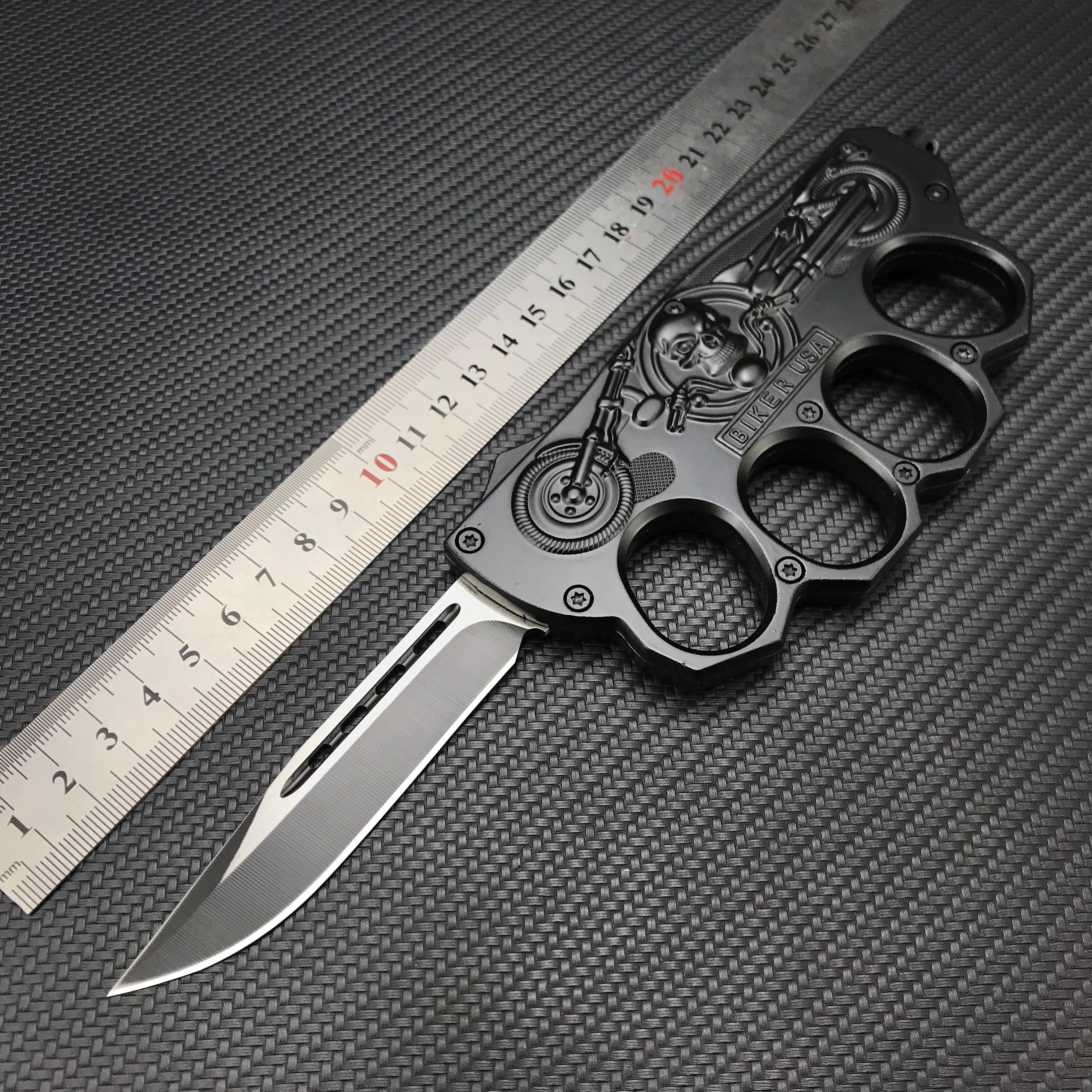 A black knuckle duster folding knife with a silver blade, featuring an embossed skull and motorcycle design with the text "BIKER USA", displayed with a metal ruler on a dark textured background for size reference.