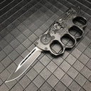 An open black folding knuckle knife featuring a silver blade with a groove, an embossed skull and motorcycle design, and "BIKER USA" text, displayed on a textured grid surface.