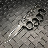 An open black folding knuckle knife featuring a silver blade with a groove, an embossed skull and motorcycle design, and "BIKER USA" text, displayed on a textured grid surface.