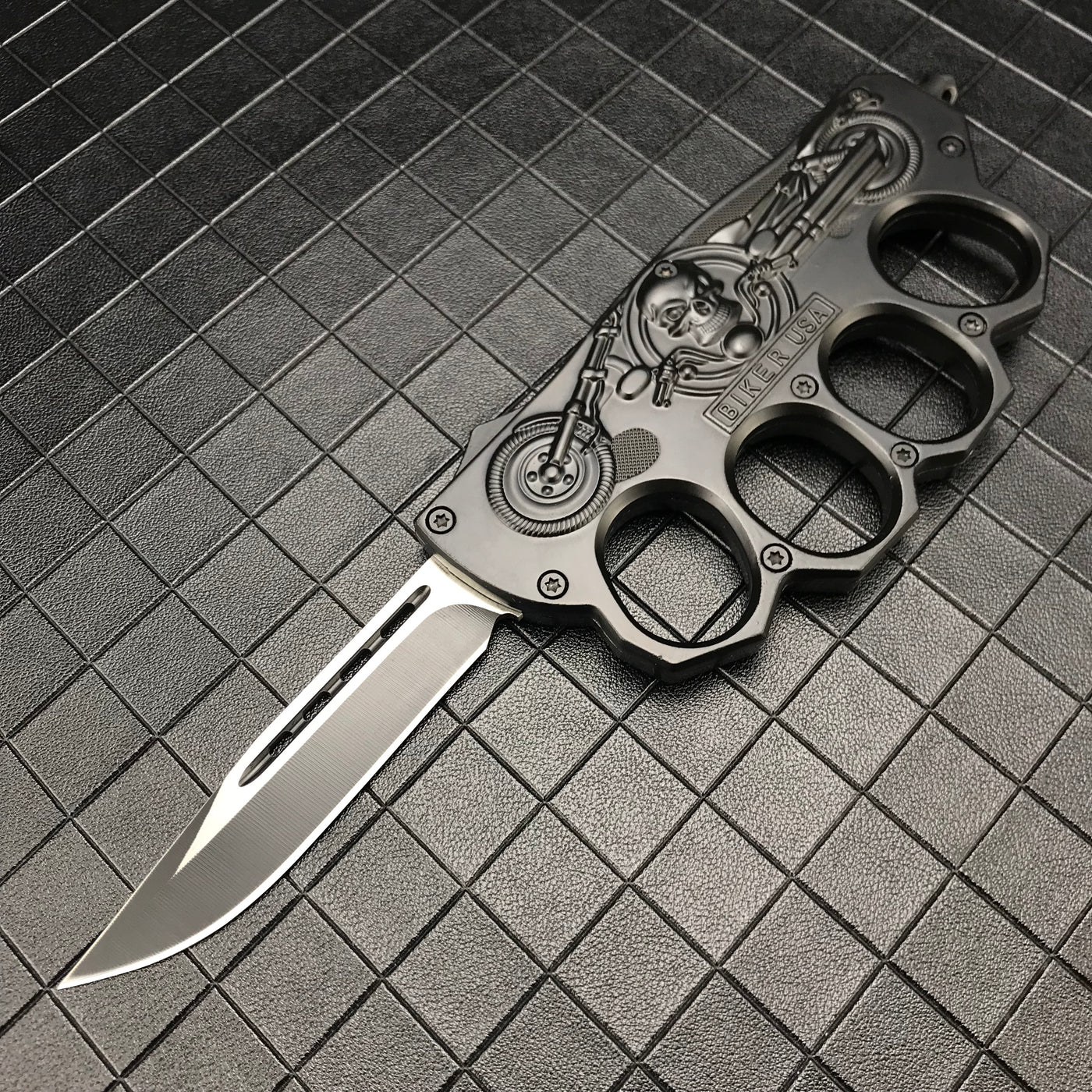 An open black folding knuckle knife featuring a silver blade with a groove, an embossed skull and motorcycle design, and "BIKER USA" text, displayed on a textured grid surface.