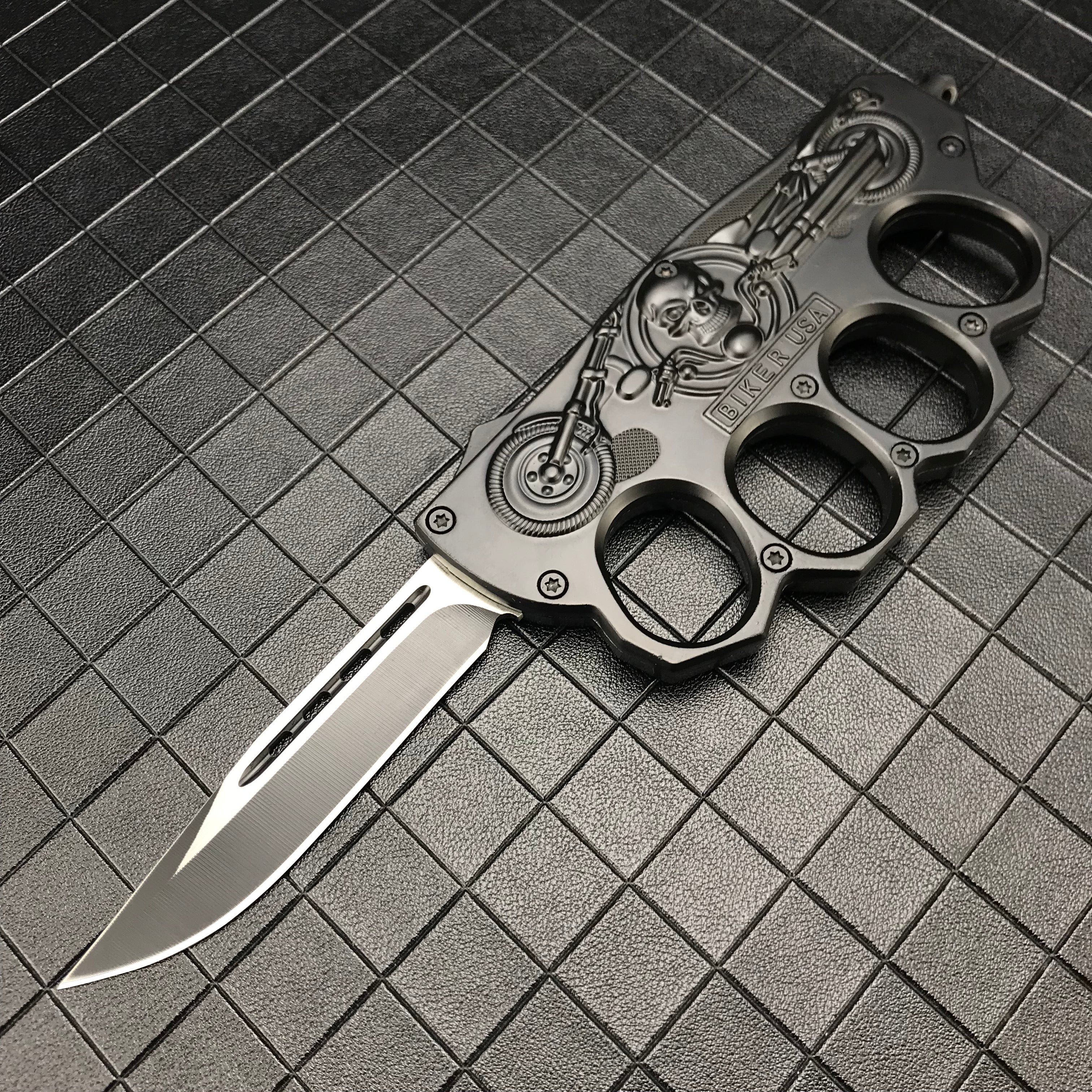An open black folding knuckle knife featuring a silver blade with a groove, an embossed skull and motorcycle design, and "BIKER USA" text, displayed on a textured grid surface.