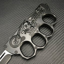 A close-up of a black knuckle duster folding knife handle, featuring an embossed skull, motorcycle design, and "BIKER USA" lettering, displayed on a carbon fiber textured background.