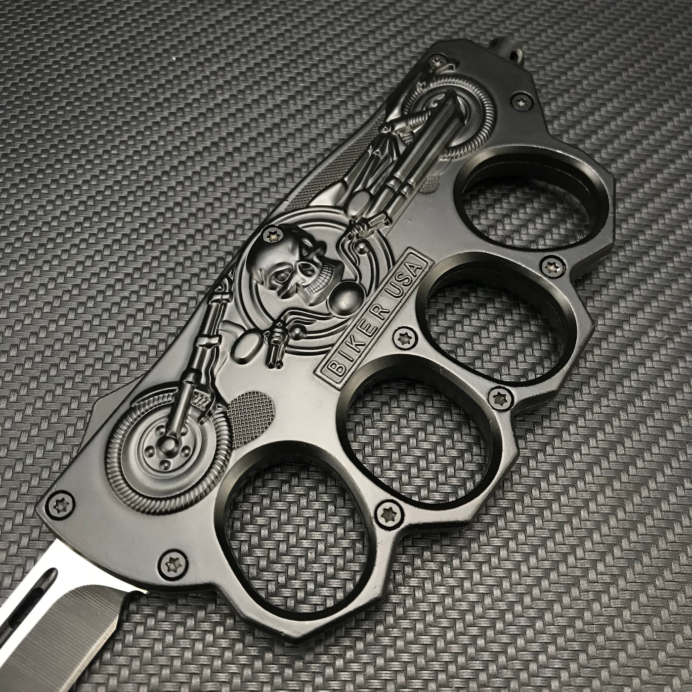 A close-up of a black knuckle duster folding knife handle, featuring an embossed skull, motorcycle design, and "BIKER USA" lettering, displayed on a carbon fiber textured background.