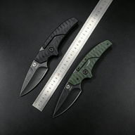 Two open tactical folding knives with black and olive green textured handles and dark blades, displayed with a metal ruler between them on a dark background for size reference.
