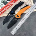 Two folding knives, one black with a blue pivot screw and one orange, featuring black blades, displayed next to a metal ruler and a red book on a grey textured surface.