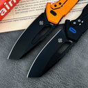 Two open tactical folding knives with black blades. The top knife features an orange handle, while the bottom knife has a black handle with a blue pivot button.