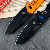 Two open tactical folding knives with black blades. The top knife features an orange handle, while the bottom knife has a black handle with a blue pivot button.