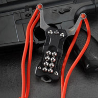 A black G10 handle EDC slingshot with a stainless steel fork, red high-elastic band, and built-in steel ball storage, displayed on a dark tactical firearm background.