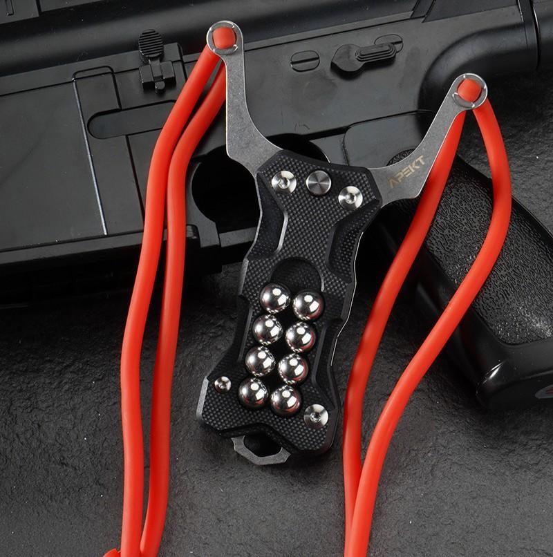 A black G10 handle EDC slingshot with a stainless steel fork, red high-elastic band, and built-in steel ball storage, displayed on a dark tactical firearm background.