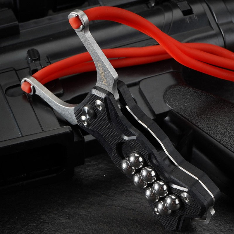 A black G10 handle EDC slingshot with a stainless steel fork, red high-elastic band, and steel ball storage, displayed on a dark tactical firearm background.