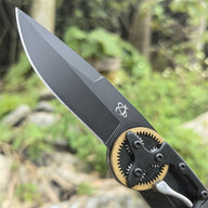 A close-up of the black blade and gold gear pivot of a folding knife, showing the engraved logo against an outdoor forest background.