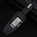 A sheathed folding knife with a black textured handle and silver hardware, resting inside a black hard plastic sheath equipped with a silver metal belt clip featuring a stylized logo, displayed on a dark background.