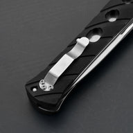 A close-up of a black folding knife handle featuring a silver pocket clip secured by three screws and circular cutouts along the handle, displayed on a dark grey background.