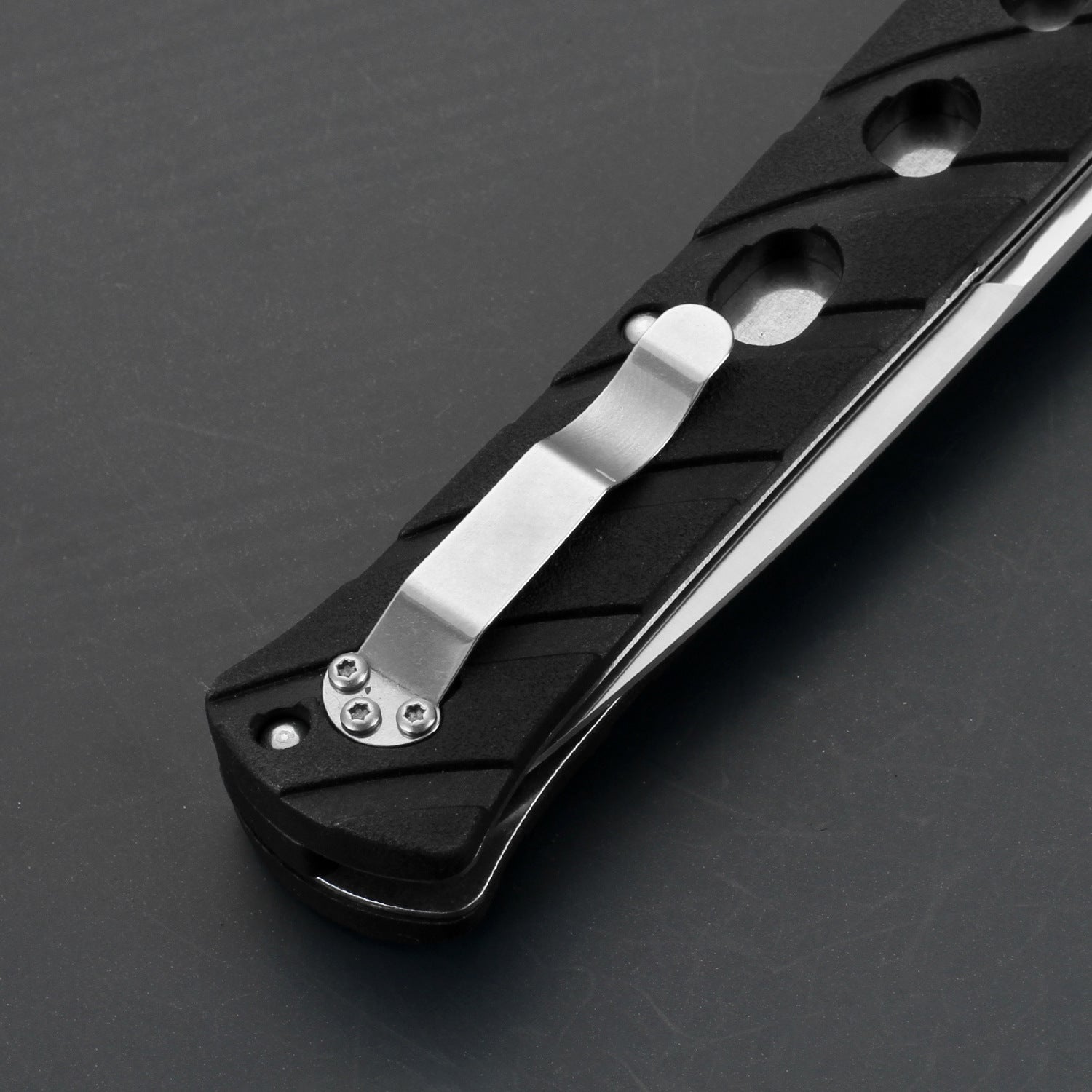 A close-up of a black folding knife handle featuring a silver pocket clip secured by three screws and circular cutouts along the handle, displayed on a dark grey background.