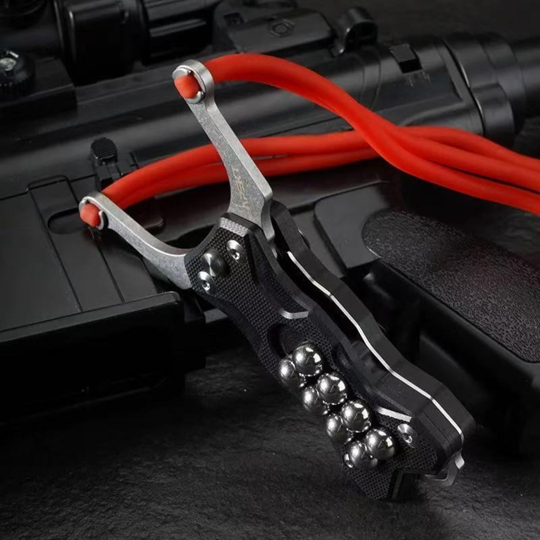 A black G10 handle EDC slingshot with a red rubber band and steel ball storage, displayed on a dark tactical gear background.