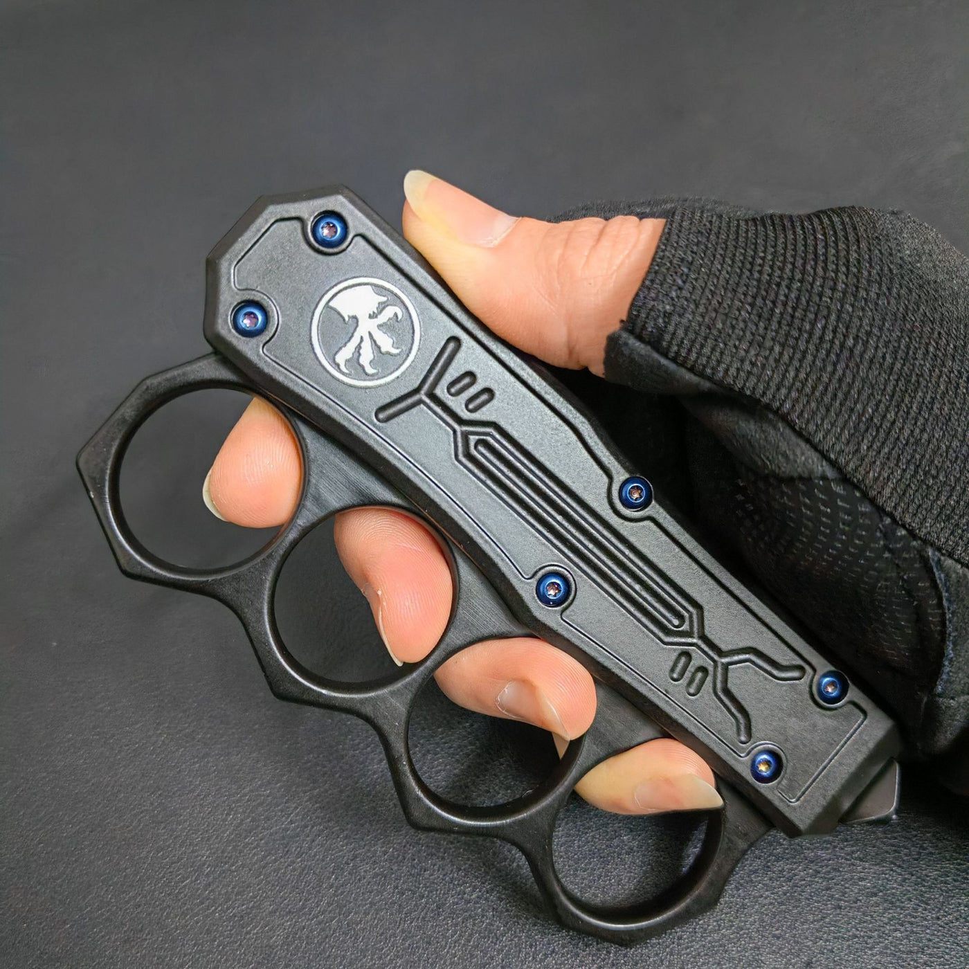 A close-up of a closed black knuckle duster OTF knife held by a hand, highlighting the white logo, blue screws, and textured handle frame.