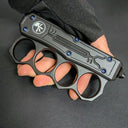 A close-up of a closed black OTF knife with a knuckle duster handle, showing the white logo, blue screws, and four finger holes, held by a hand in a black tactical glove.