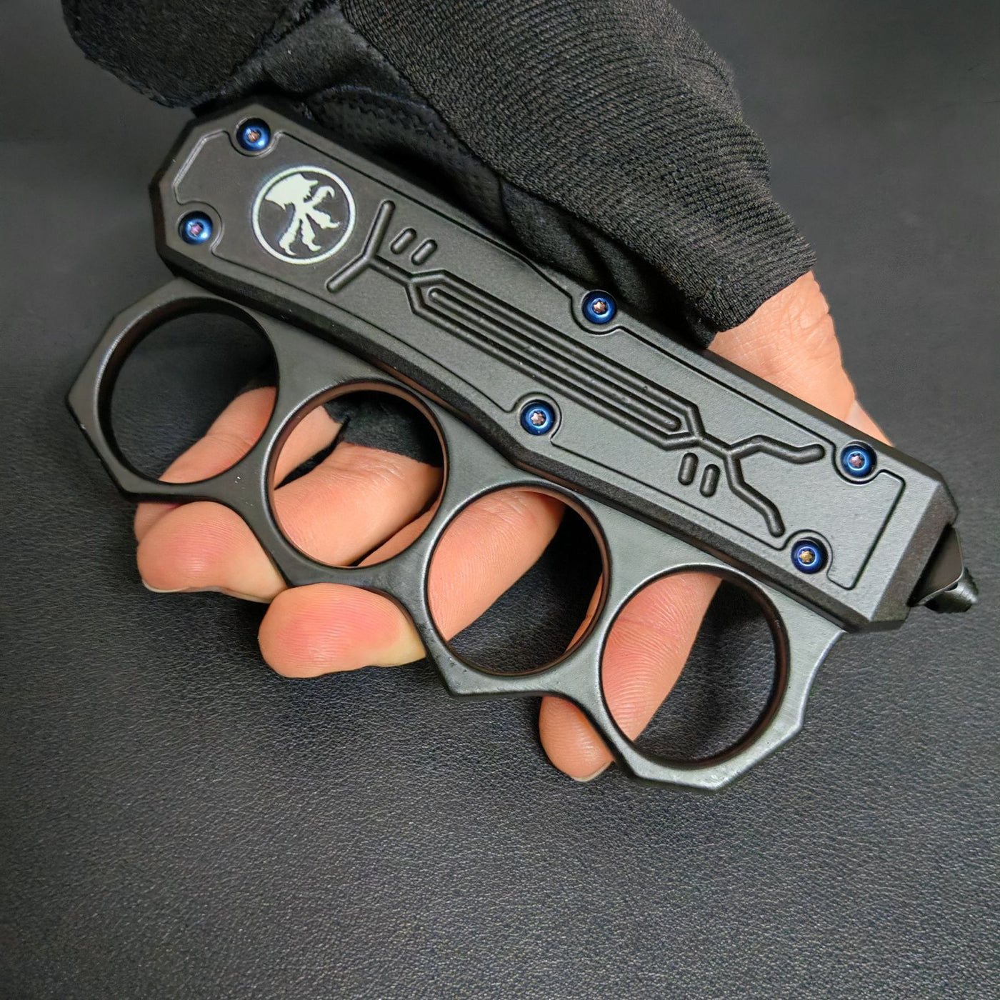 A close-up of a closed black OTF knife with a knuckle duster handle, showing the white logo, blue screws, and four finger holes, held by a hand in a black tactical glove.