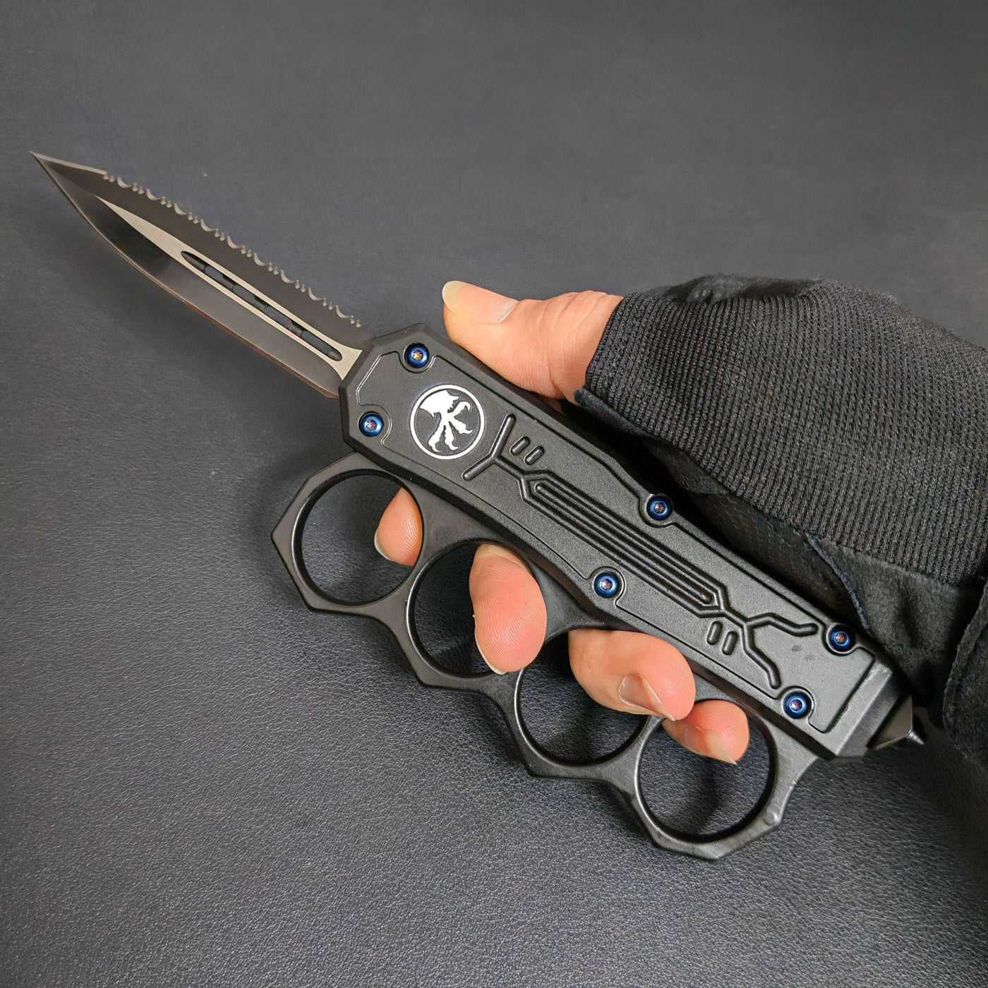 A hand wearing a tactical glove holding an open black OTF knife with a knuckle duster handle, partially serrated blade, and blue screws.