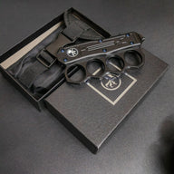 A closed black knuckle duster OTF knife resting on its packaging box and black nylon sheath, displayed on a dark surface.