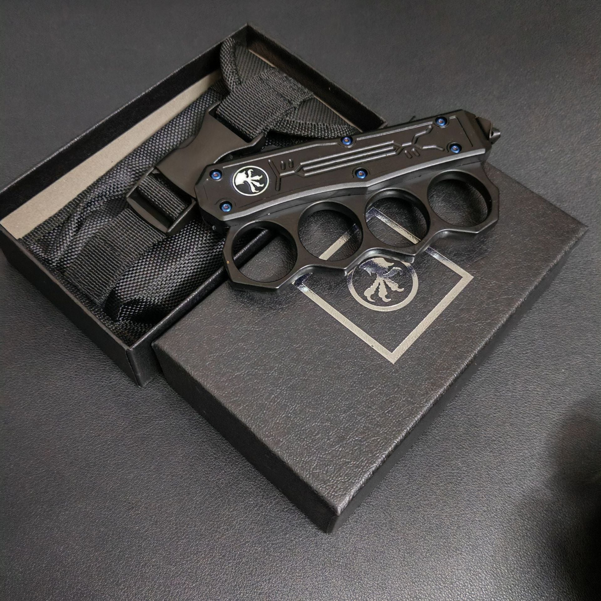 A closed black knuckle duster OTF knife resting on its packaging box and black nylon sheath, displayed on a dark surface.