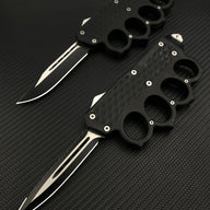 Two open black tactical OTF knives featuring knuckle duster handles with textured grips, displayed on a dark carbon fiber patterned background.