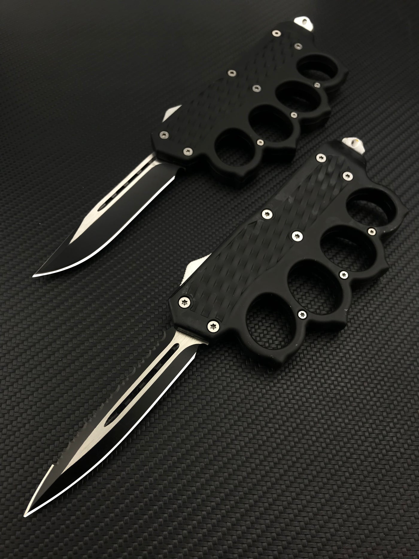 Two open black tactical OTF knives featuring knuckle duster handles with textured grips, displayed on a dark carbon fiber patterned background.