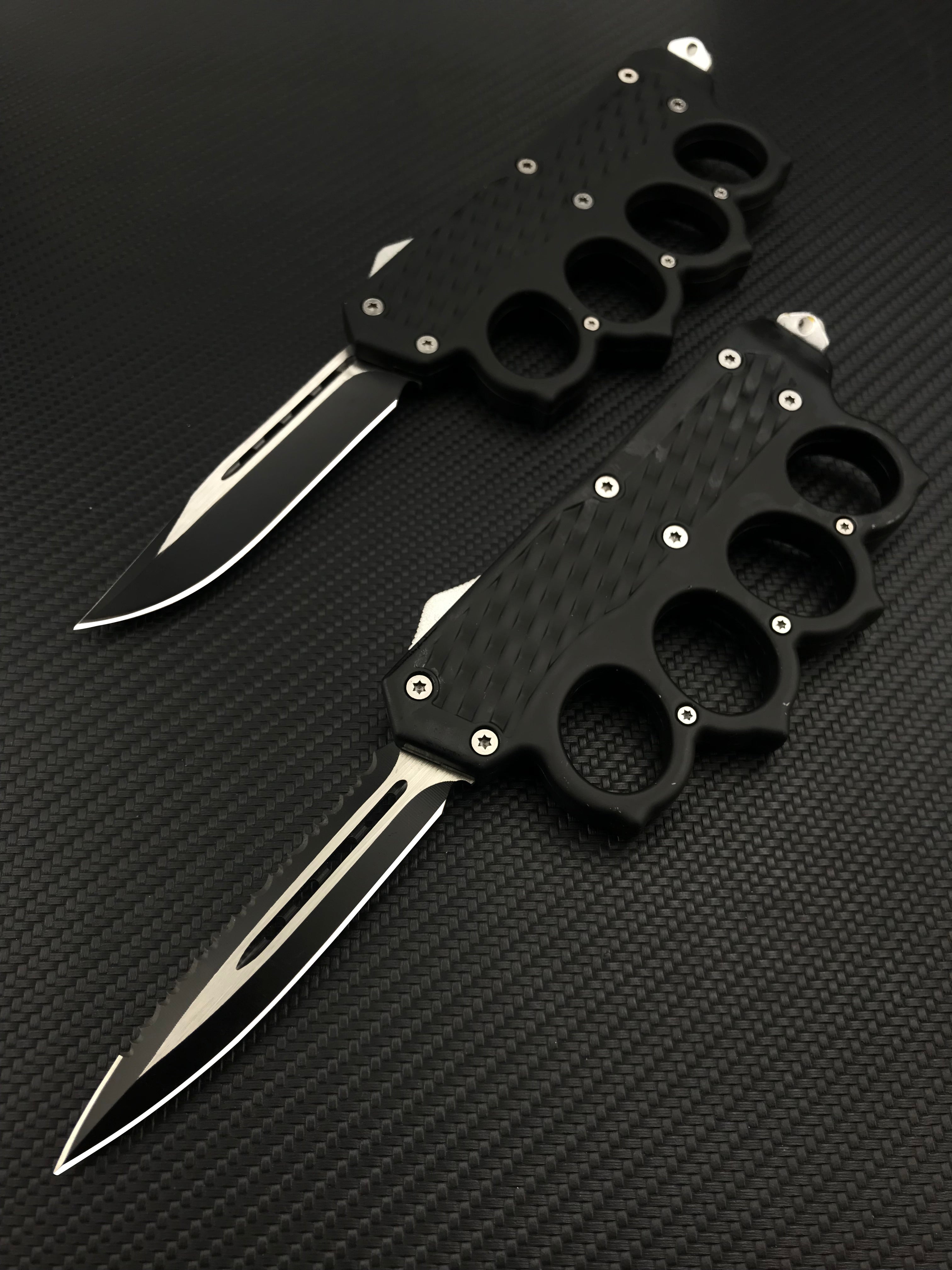 Two open black tactical OTF knives featuring knuckle duster handles with textured grips, displayed on a dark carbon fiber patterned background.