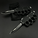 Two open black OTF knives with knuckle duster handles, one with a plain edge and one with a serrated edge, displayed on a black nylon sheath against a dark background.