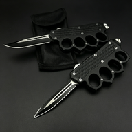 Two open black OTF knives with knuckle duster handles, one with a plain edge and one with a serrated edge, displayed on a black nylon sheath against a dark background.